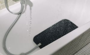 Badewanne mit Handbrause, laufendem Wasser und schwarzer Antirutschmatte auf dem Boden.