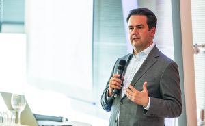 Mann im grauen Anzug hält Mikrofon und spricht vor Publikum, neben ihm ein Laptop auf einem Stehtisch.