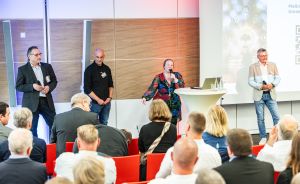 Vier Personen auf einer Bühne vor Publikum in roten Stühlen; Frau spricht ins Mikrofon, Präsentation im Hintergrund.