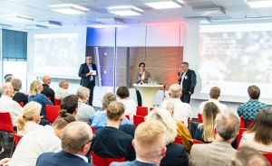 Drei Personen stehen vorne in einem Konferenzraum, dahinter sitzen Zuhörer auf roten Stühlen vor einer Leinwand.