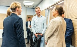 Drei Personen stehen in einem Büroflur und unterhalten sich. Ein Mann lächelt, hält eine Jacke. Im Hintergrund zwei weitere Personen.