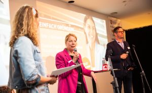 Drei Personen auf einer Bühne, eine Frau im rosa Blazer spricht ins Mikrofon, Projektionswand mit "Einfach Gründen!"