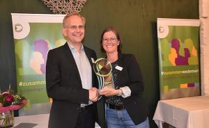 Zwei Personen halten gemeinsam eine Trophäe. Im Hintergrund Werbebanner mit Hashtag #zusammen.