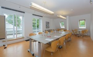 Heller Konferenzraum mit langen Tischen, Holzstühlen, Wassergläsern, großen Fenstern und Fluoreszenzbeleuchtung.