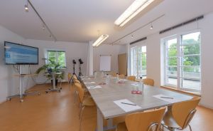 Heller Konferenzraum mit langem Tisch, Holzstühlen, Flipchart, TV-Bildschirm und großen Fenstern mit Grünblick.