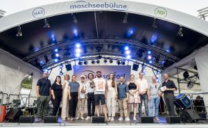 Gruppe von ca. 15 Personen auf der Maschseebühne Hannover, dahinter Bühnenbeleuchtung und „Maschseefest"-Banner.