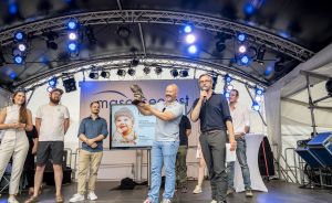 Mehrere Personen auf einer beleuchteten Bühne; zwei Männer halten eine Trophäe, im Hintergrund eine Leinwand.