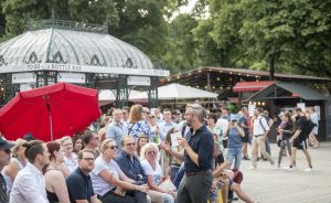 Ein Mann spricht vor einer Menschengruppe im Freien, im Hintergrund ein Pavillon mit "Bottle Bar"-Schild.