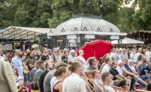 Menschenmenge sitzt und steht im Freien vor einem Pavillon mit Glaskuppel, roter Sonnenschirm, Bäume im Hintergrund.