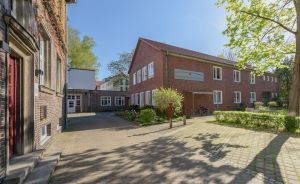 Gepflasterter Innenhof mit roten Backsteingebäuden, Grünflächen und Bäumen bei sonnigem Wetter.