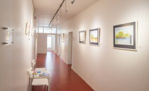 Langer Galerieflur mit rotem Boden, weißen Wänden, Deckenspots und gerahmten Landschaftsgemälden. Weißer Stuhl links.