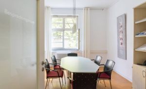 Besprechungsraum mit ovalem Tisch, sechs Stühlen, einem Fenster mit Vorhängen und einem Bücherregal.