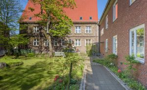 Backsteingebäude mit rotem Dach, gepflegter Garten mit Rasenfläche, Gartenweg, Bäume und Blumenbeete bei Sonnenschein.