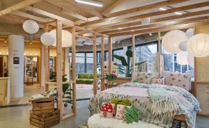 Helles Ladengeschäft mit Holzrahmen, weißen Lampions, einem dekorierten Bett, Holzkisten und Zimmerpflanzen.