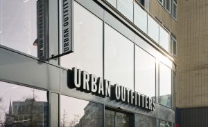 Außenansicht eines Urban Outfitters-Geschäfts mit Glasfassade, großem Schriftzug und Flaggenschild an einem Stadtgebäude.