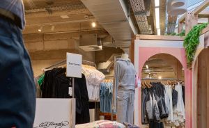 Ed Hardy Kleidungsabteilung mit gefalteten Shirts, Markenaufsteller, Kleiderständern und Schaufensterpuppe im Hintergrund.