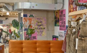 Innenraum eines Geschäfts mit orangefarbenem Sofa, blauem Tisch, Postern, Pflanzen und hängenden Jeans auf einem Kleiderständer.
