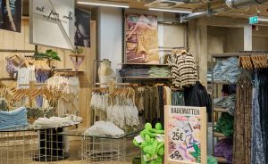 Innenansicht eines Bekleidungsgeschäfts mit Holztischen, Badetüchern, Preisschild „Badematten 25€" und Kleidung auf Ständern.