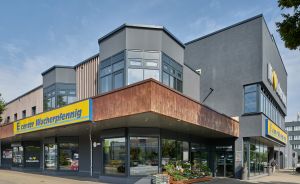 Außenansicht eines E-Center-Supermarkts mit gelbem Schriftzug „Wucherpfennig", grauer Fassade und Blumendisplay vor dem Eingang.
