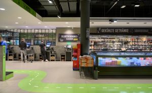 Supermarkt mit Selbstbedienungskassen, Getränkekühlregal und grüner Wegemarkierung auf dem Boden.