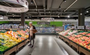 Supermarkt-Obst- und Gemüseabteilung mit bunten Auslagen, einer Person in Bewegung und Schildern auf Deutsch.