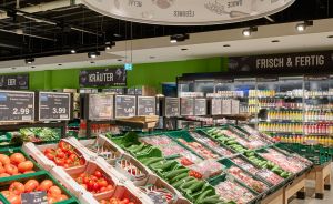 Gemüseabteilung eines Supermarkts mit Gurken, Tomaten und Paprika in grünen Kisten. Hängeschild mit "Sebze/Fruit/Fruta".