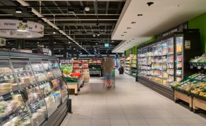 Supermarkt-Innenraum mit Gemüse- und Kühlregalen, zwei Kunden im Gang, grüne Wandgestaltung, helle Deckenbeleuchtung.