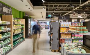 Supermarkt-Gang mit Person in Bewegung, links Kräuter-Regal, rechts Konserven und Müsli-Produkte, modernes Beleuchtungssystem.