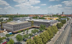 Luftaufnahme einer Stadt mit mehrstöckigen Gebäuden, breiter Straße, Bäumen und bewölktem Himmel. Rechts ein gelbes POCO-Gebäude.