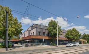 Moderne Gebäude mit Holzfassade an einer Stadtstraße mit Straßenbahngleisen, Bäumen und parkenden Autos.
