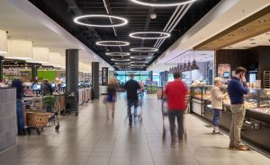 Moderner Supermarkt mit runden LED-Deckenleuchten, Einkaufende auf Fliesenboden, links Kassen, rechts Bäckerei-Theke.