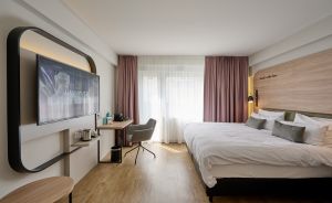 Hotelzimmer mit Doppelbett, Holzverkleidung, Wandmontiertem TV, Schreibtisch, grauem Stuhl und rosafarbenen Vorhängen.