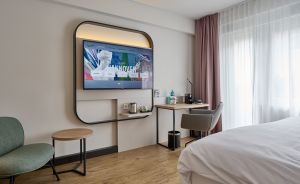 Modernes Hotelzimmer mit TV, Schreibtisch, Sessel, Bett und rosa Vorhängen vor einem Fenster.