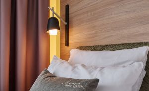 Hotelzimmer mit weißen Kissen, grauem Zierkissen, Wandlampe und Holzkopfteil mit Schriftzug „travel. work. Live."