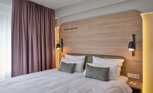 Hotelzimmer mit Doppelbett, hellem Holzkopfteil, Wandleuchten, grauen Kissen und rosafarbenen Vorhängen.