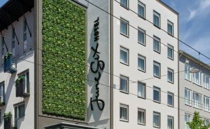 Modernes Stadtgebäude mit begrünter Fassadenwand, Glaseingang, Fahrrädern und Fußgängern auf dem Gehweg.