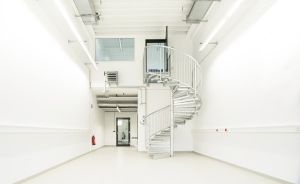 Weißer Industrieraum mit silberner Wendeltreppe, Oberlicht, Neonröhren, rotem Feuerlöscher und grauer Tür.