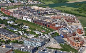 Luftaufnahme eines Stadtgebiets mit Baustellen, Kränen, mehrstöckigen Gebäuden, Grünflächen und Feldern im Hintergrund.