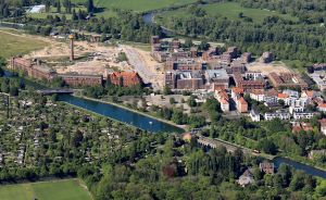 Luftaufnahme einer Stadt mit Kanal, roten Backsteingebäuden, Wohnhäusern, Grünanlagen und einer Baustelle.