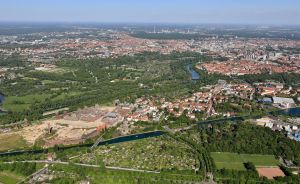 Luftaufnahme einer Stadt mit roten Ziegeldächern, Fluss, Grünanlagen und einem großen historischen Gebäudekomplex im Vordergrund.