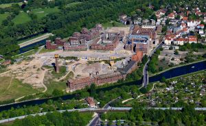 Luftaufnahme einer Stadt mit Industriegebäuden, Baustelle, Kanal, Wohnhäusern und umgebenden Grünflächen.