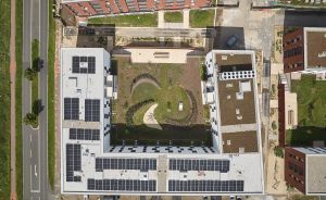 Luftaufnahme eines U-förmigen Gebäudes mit Solarpanelen auf dem Dach und begrüntem Innenhof mit geschwungenen Wegen.