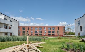 Moderner Wohnkomplex mit rotem Backsteingebäude, grüner Rasenfläche, Holzspielstruktur im Sandbereich und blauem Himmel.