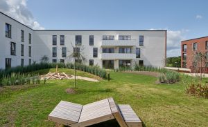 Modernes weißes Mehrfamilienhaus mit Grünanlage, Liegestühlen aus Holz und bepflanzten Beeten im Innenhof.