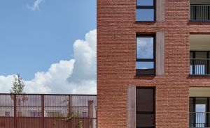 Rotes Backsteingebäude mit vier Stockwerken, Balkon, Metallgitter-Zaun und Sträuchern vor blauem Himmel.