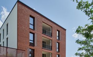 Modernes Wohngebäude mit roter Backsteinfassade, Balkonen, weißem Anbau, Begrünung und Spielplatz bei blauem Himmel.