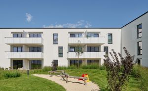Weißes Mehrfamilienhaus mit Balkonen, davor Kinderspielplatz mit Sandkasten und Rutsche, umgeben von Rasenfläche.