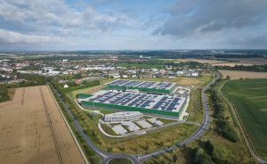 Luftaufnahme eines großen Industriegebäudes mit Solarpanelen auf dem Dach, umgeben von Feldern und einer Kleinstadt.