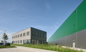 Modernes Industriegebäude mit grüner und grauer Fassade neben zweigeschossigem Bürogebäude, blauer Himmel, Grünflächen.