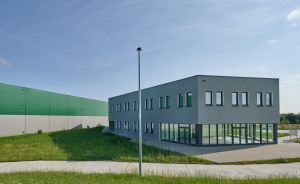 Zweigeschossiges graues Bürogebäude mit grünen Akzenten, daneben grüne Industriehalle, Grünfläche und Straßenlaterne.